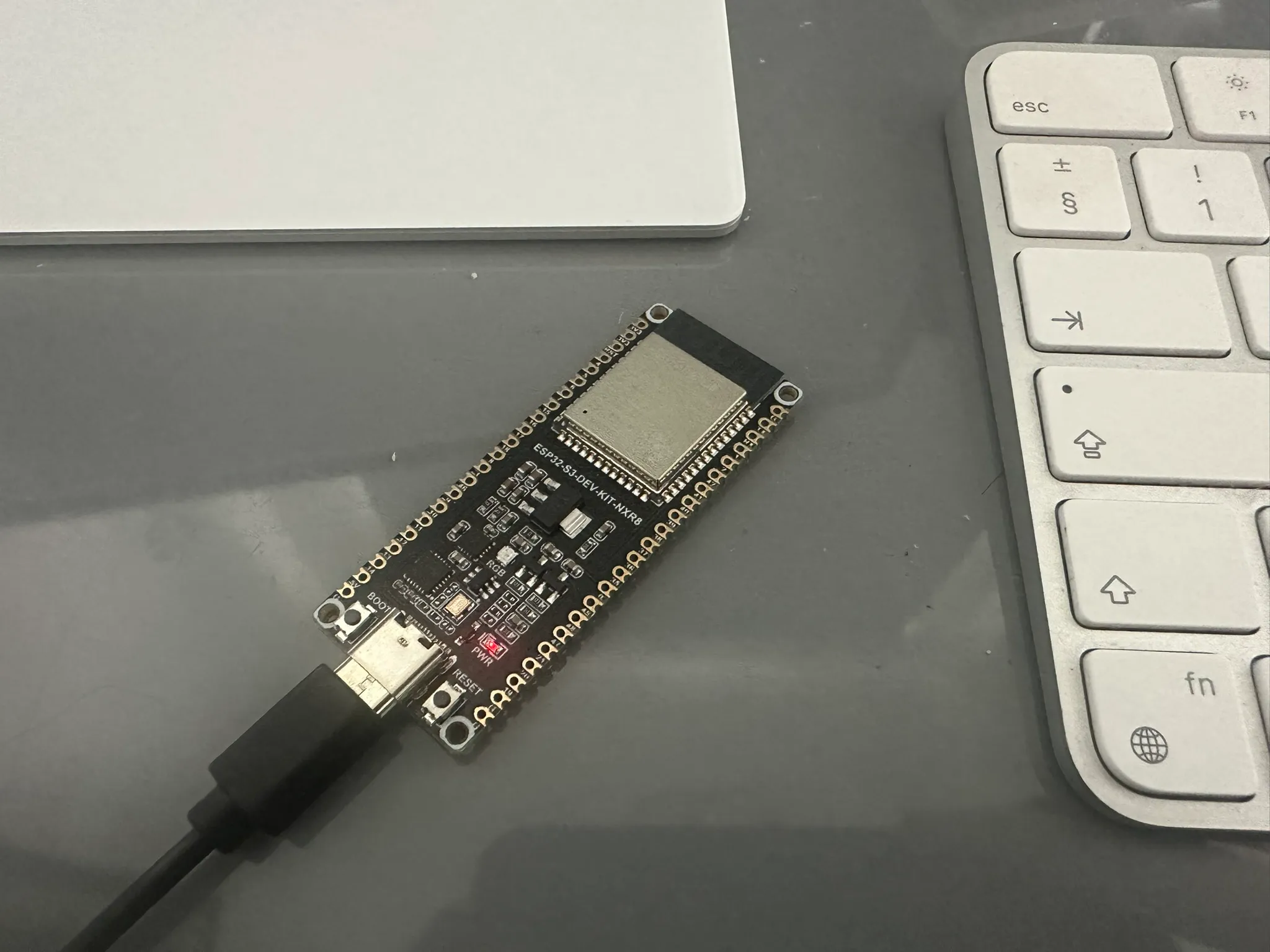 An ESP32 dev board - a small printed circuit board with a red LED lit, resting on a grey desk.