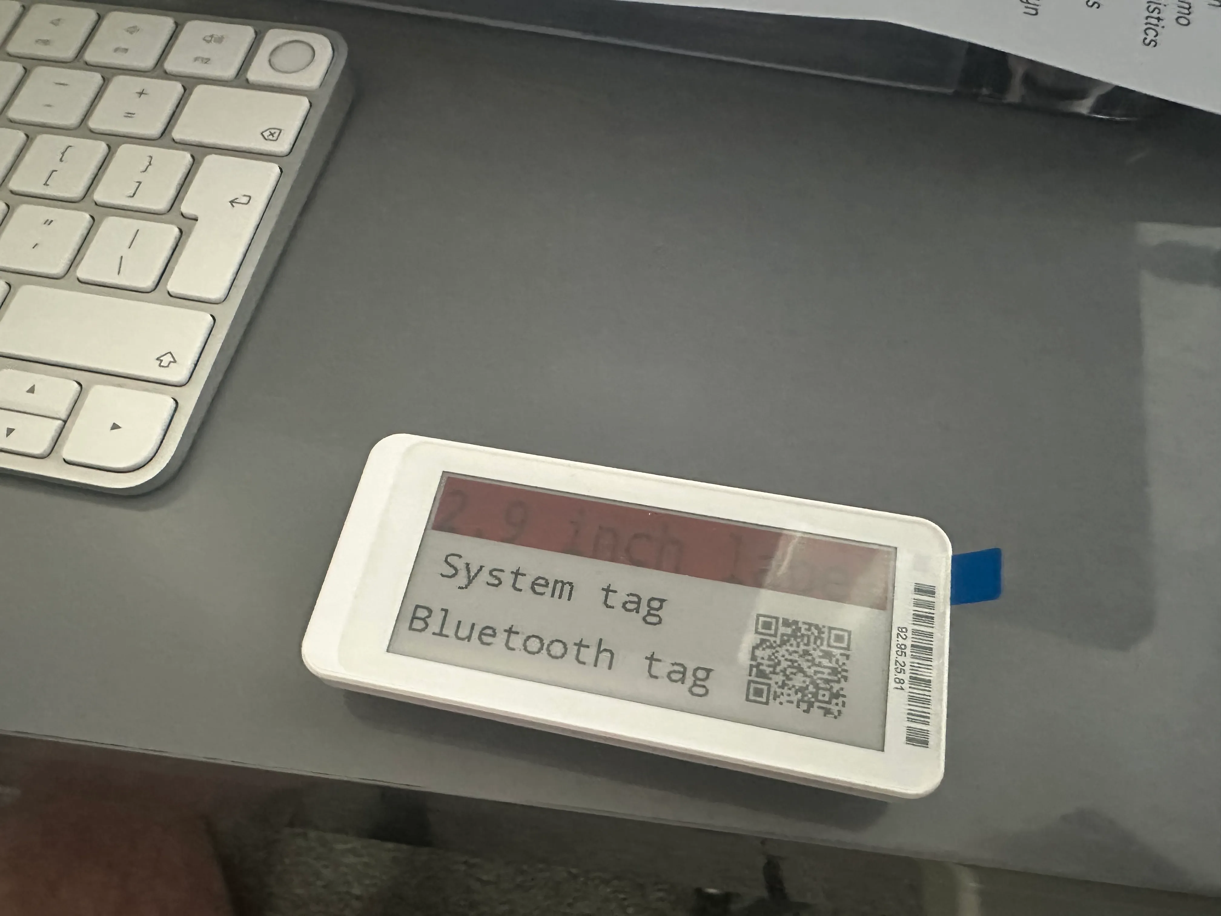 An Electronic Shelf Label sitting on a grey desk next to a Mac keyboard.